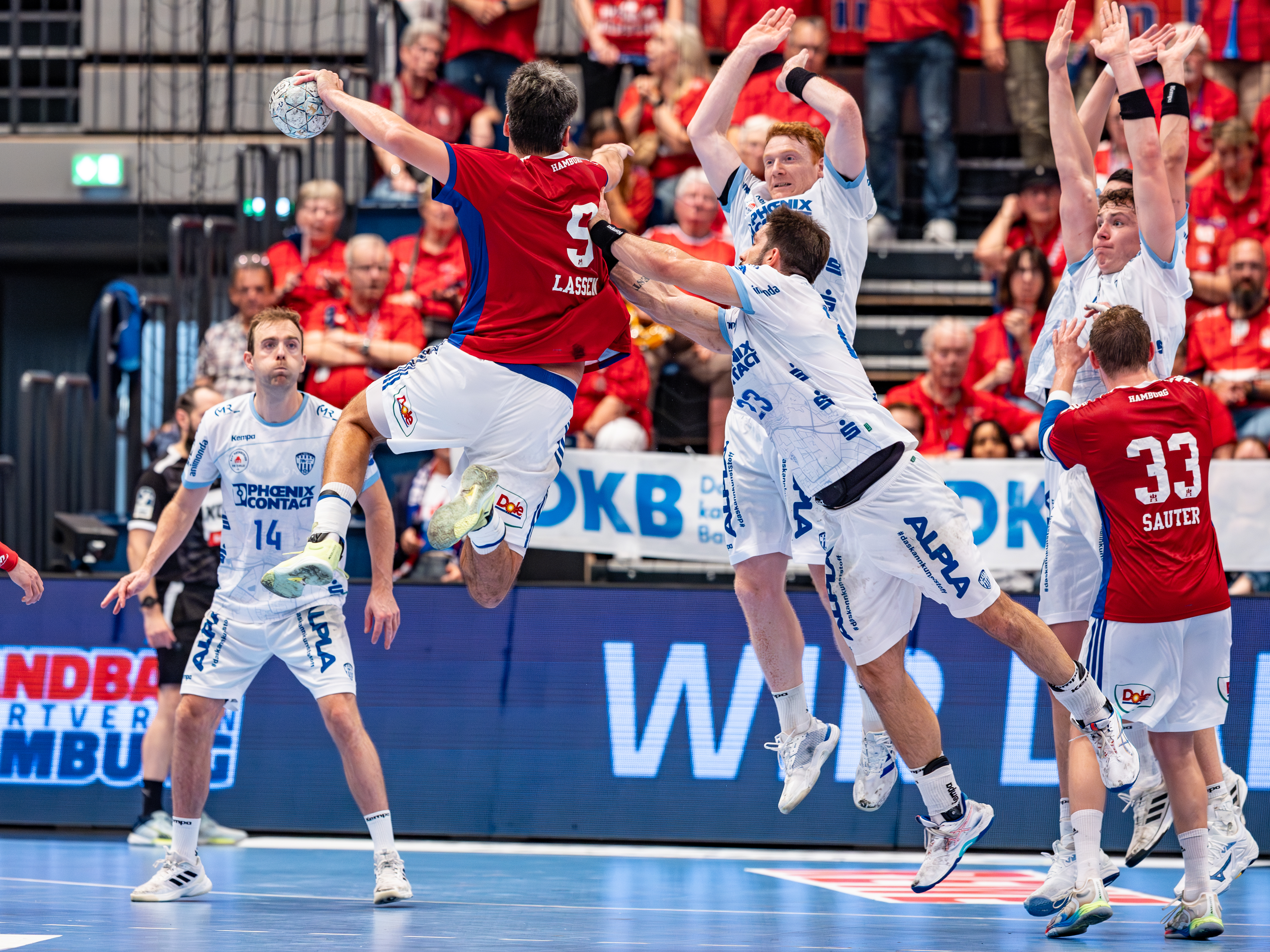 #9 Jacob Lassen (Rückraum rechts) – Handball Sport Verein Hamburg jump high and shoots at goal. #23 Tim Suton (Rückraum Mitte) – TBV Lemgo Lippe and #3 Nicolai Theilinger (Rückraum rechts) – TBV Lemgo Lippe block.